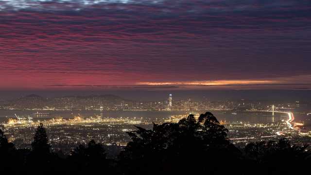 A panoramic nighttime view of a cityscape with illuminated buildings, a bridge, and a vibrant red and purple sky.