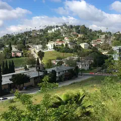 A suburban area with houses on hillsides is surrounded by trees and greenery, set against a blue sky with white clouds.