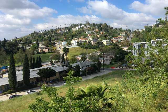 A suburban area with houses on hillsides is surrounded by trees and greenery, set against a blue sky with white clouds.
