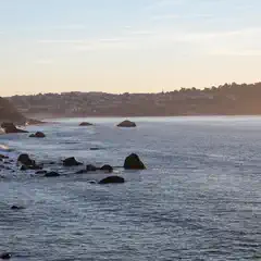 A rocky shoreline at sunset with a calm sea and distant houses on the horizon.