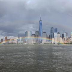 A rainbow arcs over a river in front of a city skyline with tall buildings.