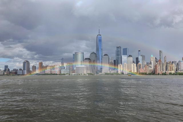 A rainbow arcs over a river in front of a city skyline with tall buildings.