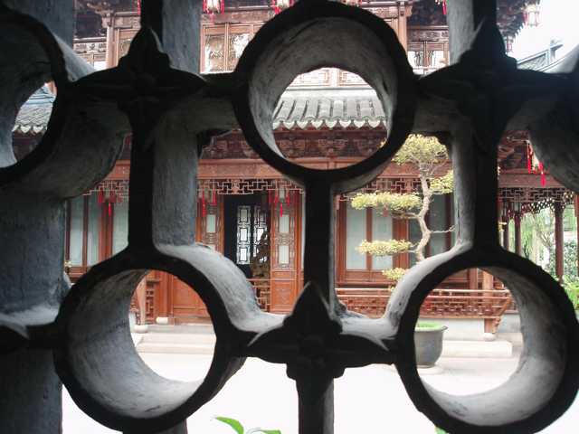 A traditional Chinese-style building with intricately carved wooden beams and lattice windows, viewed through a circular metal gate featuring similar designs.