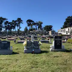 A cemetery with numerous headstones, some adorned with flowers, set against a backdrop of trees and a clear sky.