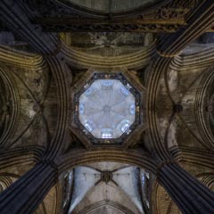 A grand, ornate ceiling with stone vaulting and ribbed arches, featuring a central octagonal lantern with stained glass windows, illuminated by soft light.