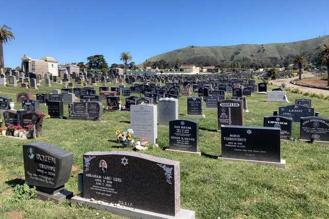 A cemetery contains numerous dark-colored headstones on a grassy hillside beneath a clear blue sky.