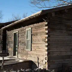 A weathered wooden cabin with a green door and window frames, set against a clear sky.