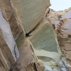 A rocky cliff face with jagged edges, showing layers and cracks in various shades of brown, tan, and green.