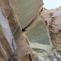 A rocky cliff face with jagged edges, showing layers and cracks in various shades of brown, tan, and green.