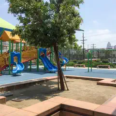 An urban park features a playground structure with yellow and red metal bars, blue plastic slides, and a green canopy.