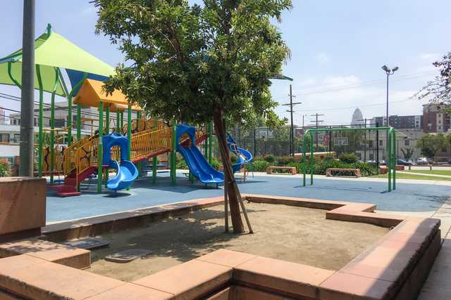 An urban park features a playground structure with yellow and red metal bars, blue plastic slides, and a green canopy.