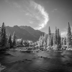 A serene black-and-white landscape featuring a flowing river, dense evergreen forest, distant mountains, and a person sitting contemplatively on a rock.