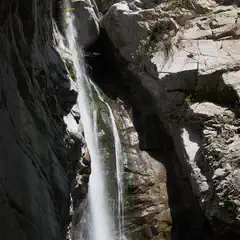 A waterfall cascading down rocky cliffs into a stream, with sunlight filtering through the canyon and illuminating the water.