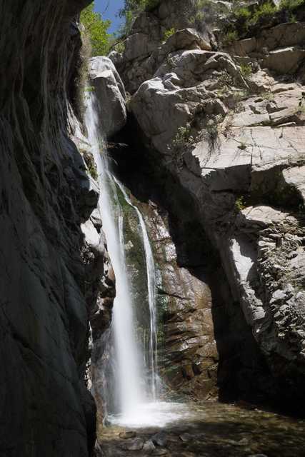 A waterfall cascading down rocky cliffs into a stream, with sunlight filtering through the canyon and illuminating the water.