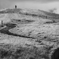 A person standing on top of a grassy hill with a fence and steps leading up to it, under a cloudy sky.