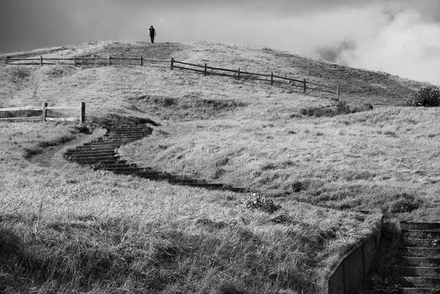 A person standing on top of a grassy hill with a fence and steps leading up to it, under a cloudy sky.
