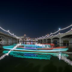 A boat decorated with colorful lights is moving on a river at night, passing under a bridge adorned with festive string lights.