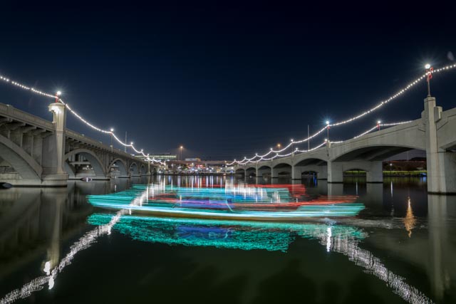 A boat decorated with colorful lights is moving on a river at night, passing under a bridge adorned with festive string lights.