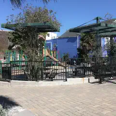 A playground structure with a slide stands behind a black metal fence and two green shaded pavilions with benches.