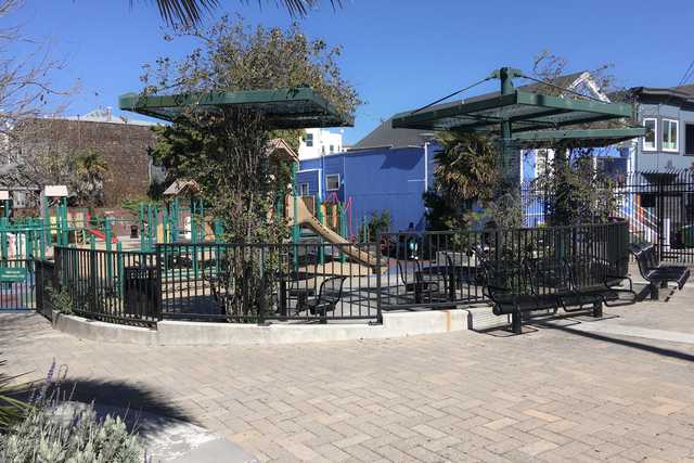 A playground structure with a slide stands behind a black metal fence and two green shaded pavilions with benches.