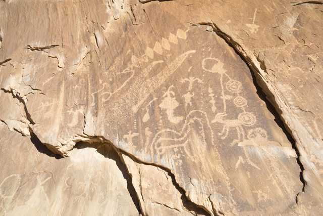 Ancient petroglyphs etched into light brown rock face, depicting various figures and symbols.