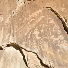 Ancient petroglyphs etched into light brown rock face, depicting various figures and symbols.