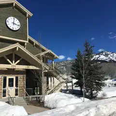 A two-story building with a clock tower and staircase, surrounded by snow-covered ground and trees, with mountains in the background.