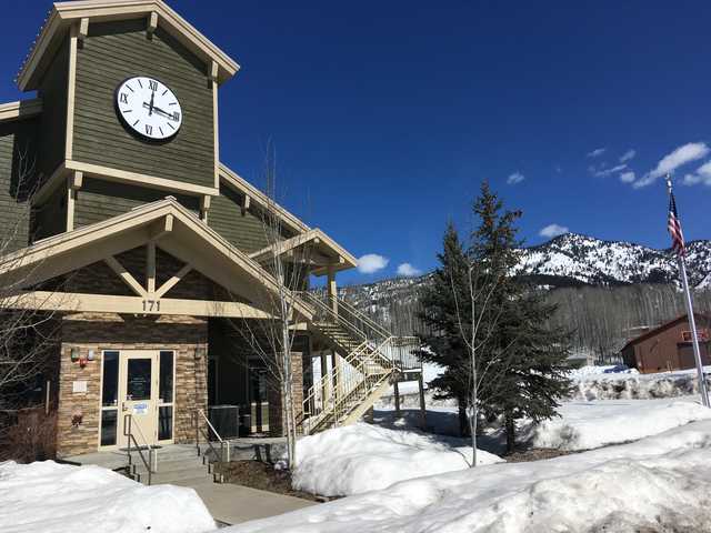 A two-story building with a clock tower and staircase, surrounded by snow-covered ground and trees, with mountains in the background.