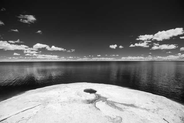 A large rock formation juts out into a calm body of water under a partly cloudy sky.