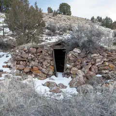 A small stone structure with a dark opening, surrounded by snow and sparse vegetation on a rocky hillside.