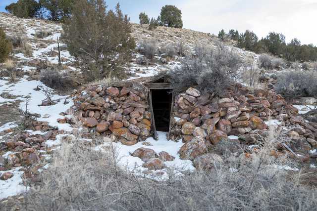 A small stone structure with a dark opening, surrounded by snow and sparse vegetation on a rocky hillside.