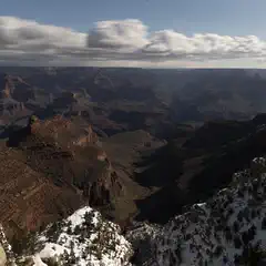 A snow-covered cliff overlooking a vast canyon with layered rock formations under a partly cloudy sky.