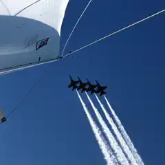 A sailboat's mast is seen from below, with four jets flying above it against a blue sky.