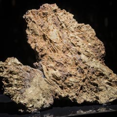 A rough, irregularly shaped gold nugget with intricate textures and a lustrous, metallic surface, displayed against a dark background.
