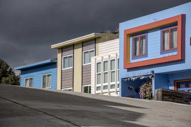 A row of colorful houses on a hillside, with dark clouds above and a concrete road below.