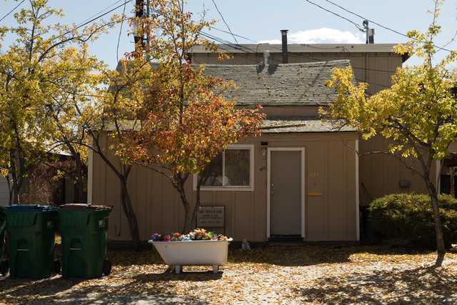 A small, brown building with a gray roof stands amidst trees and a bathtub filled with flowers in front of it.