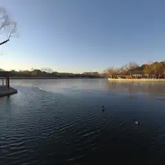 A large lake is surrounded by trees and a walkway with buildings along its edge.