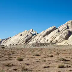 A rocky mountain range with sparse desert vegetation in the foreground and clear blue sky above.
