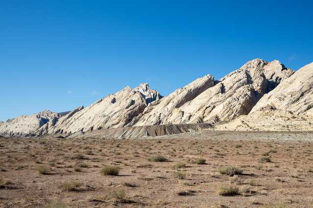 A rocky mountain range with sparse desert vegetation in the foreground and clear blue sky above.