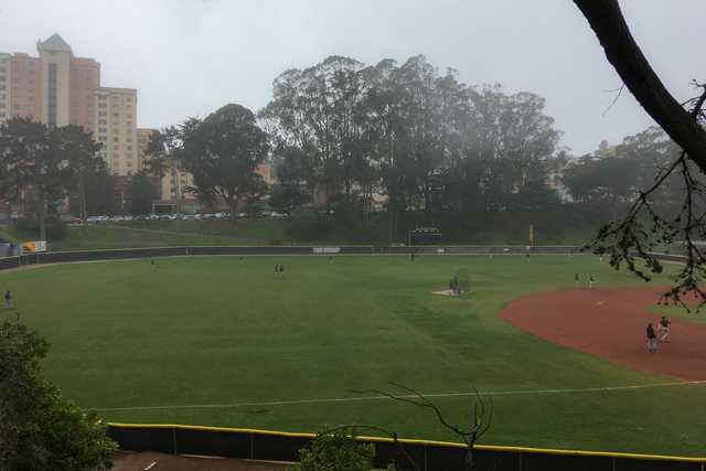 A baseball field on an overcast day with a few players visible and trees surrounding the perimeter.