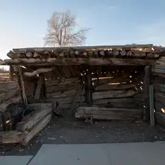 A rustic wooden structure with a sloping roof and visible gaps between the logs, on dirt ground next to a concrete path.