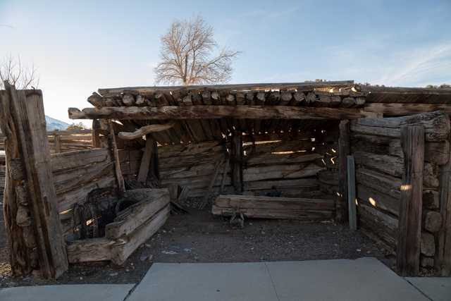 A rustic wooden structure with a sloping roof and visible gaps between the logs, on dirt ground next to a concrete path.
