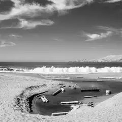 A sandy beach with a small tidal pool filled with driftwood. Waves crash in the background and mountains are visible in the distance.