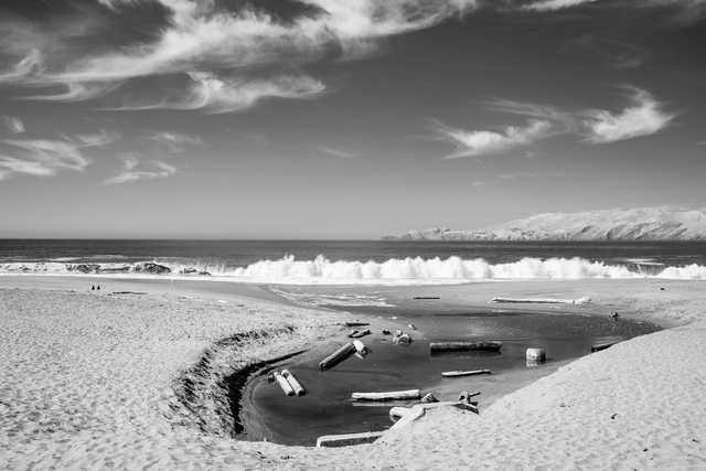 A sandy beach with a small tidal pool filled with driftwood. Waves crash in the background and mountains are visible in the distance.
