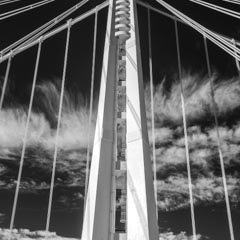 A large suspension bridge with multiple white cables and a tower visible from below on a cloudy day.