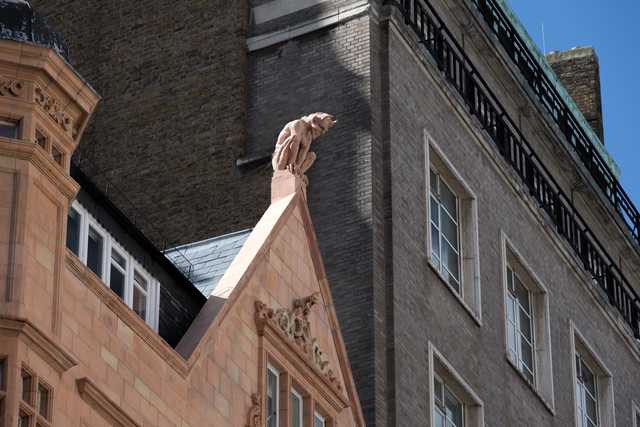 A stone gargoyle perches atop the triangular gable of a terracotta-colored building, set against a dark brick facade with multi-paned windows.