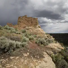 A stone ruin sits atop a rocky cliff surrounded by sparse vegetation under a cloudy sky.