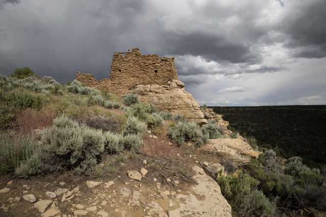 A stone ruin sits atop a rocky cliff surrounded by sparse vegetation under a cloudy sky.