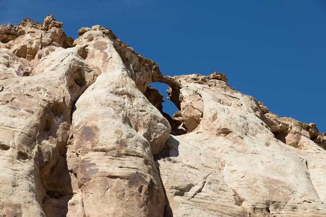 An arch-shaped rock formation is visible through a gap in a large, light-colored rocky structure against a clear blue sky.