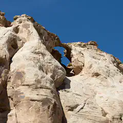 An arch-shaped rock formation is visible through a gap in a large, light-colored rocky structure against a clear blue sky.